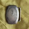 Metal belt buckle with an ornate engraved border and a central J monogram on yellow fabric.