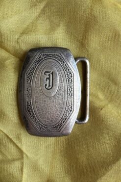 Metal belt buckle with an ornate engraved border and a central J monogram on yellow fabric.