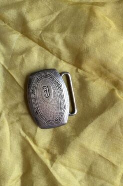 Metal belt buckle with ornate oval pattern and a raised 'J' monogram on yellow fabric backdrop.