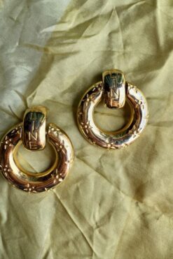 Two gold hoop earrings with star-shaped engravings resting on pale green fabric.