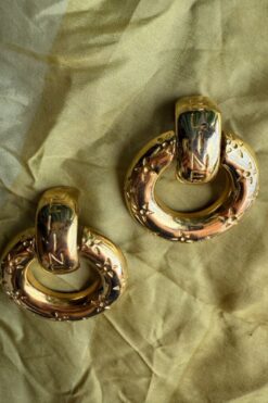 Two gold-toned hoop earrings with textured detailing on a pale fabric background.