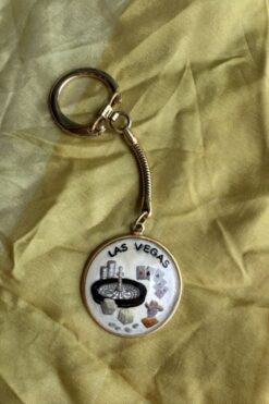 Keychain with a circular Las Vegas pendant showing a roulette wheel, dice, cards, and chips on a yellow fabric background.