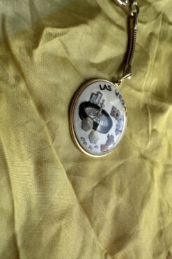 Circular gold-tone pendant with a Las Vegas themed collage (roulette wheel, chips, casino objects) on yellow fabric, edge labeled 'LAS VEGAS'