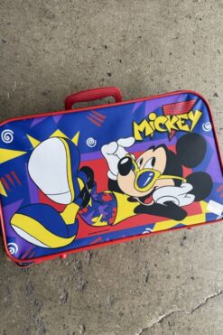 Bright blue Mickey Mouse suitcase with yellow shoes on a concrete surface nearby.