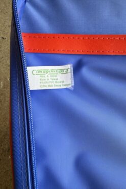 Close-up of a blue backpack with a red horizontal strap and a small tag that reads 'Imaginings' and 'The Walt Disney Company' inside the pocket area.