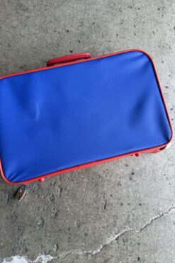 Blue hard-shell suitcase with red trim lying on a concrete floor; zipper visible at corner.