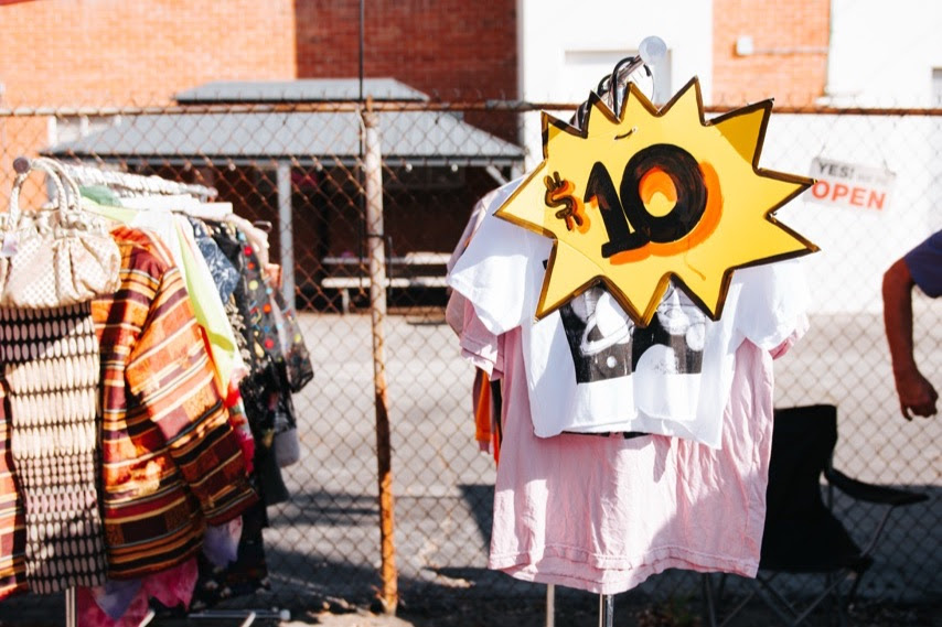 Outdoor clothing rack at a market with a bright yellow starburst sign showing '$10' for items on sale.