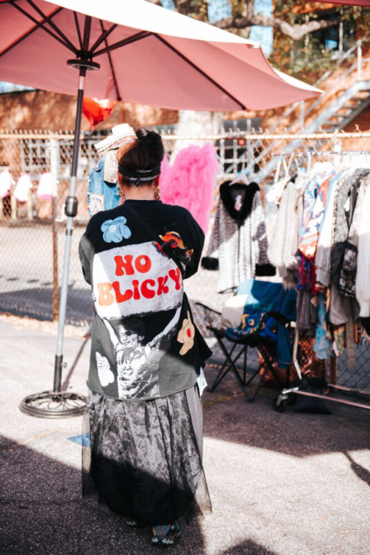Person with dark hair in a bun wearing a black graphic tee with red lettering, standing near clothing racks under a pink umbrella at an outdoor market.
