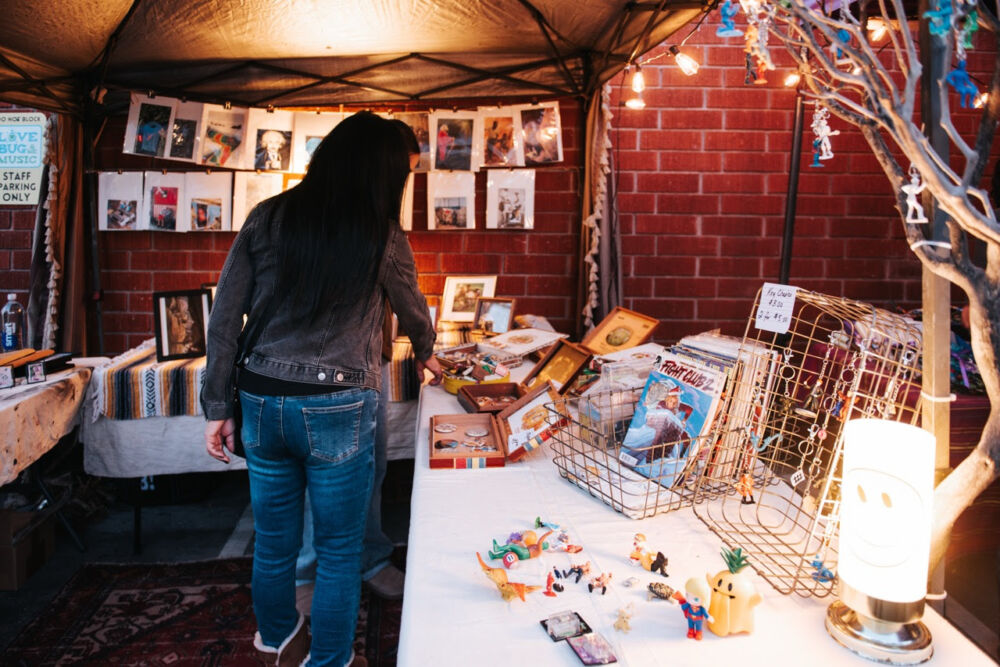 Back view of a woman in a denim jacket browsing a market stall filled with photos, frames, and trinkets under warm string lights.