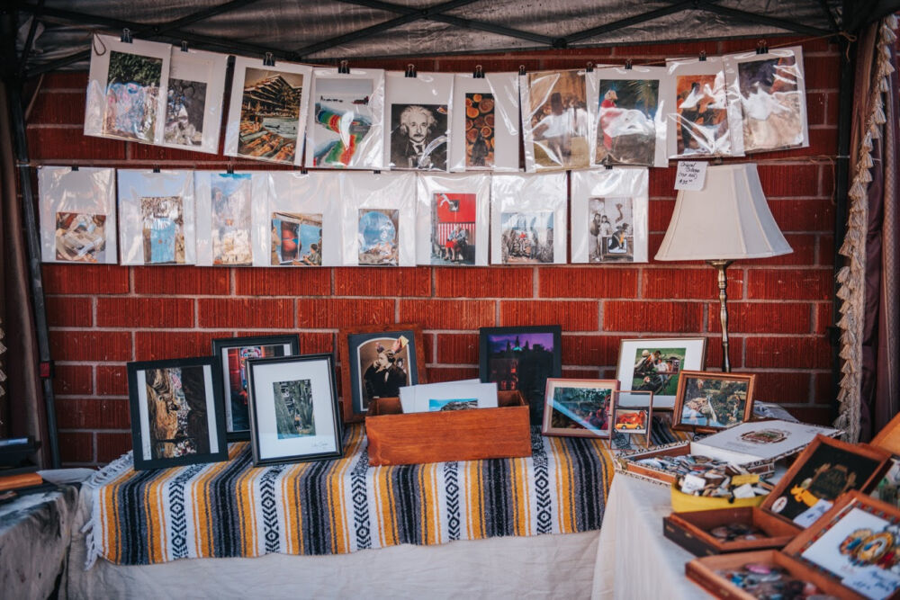 Outdoor market stall displaying framed photos and prints on a striped tablecloth with more pictures hanging on a line above the table against a brick wall.